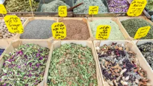 A colorful display of various dried herbs, teas, and seeds at a market stall, including labeled bins of ginger tea, flax, fennel, Arabic chai, and anis tea—highlighting diverse natural ingredients used for wellness and nutrition.