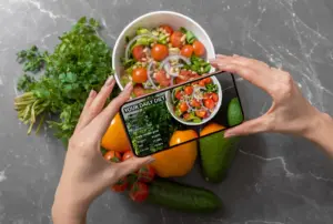 A person holds a smartphone over a fresh salad bowl, with the screen displaying a digital nutrition breakdown including protein, carbs, fats, and calories—surrounded by healthy ingredients like parsley, bell peppers, avocado, and cucumber—illustrating smart food logging and macro tracking.
