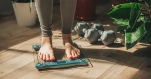 A person stands barefoot on a digital glass scale in a sunlit room, with grey dumbbells and green indoor plants nearby, representing a focus on weight tracking and healthy living—ideal for Cronometer's wellness and nutrition themes.
