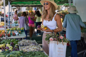 A woman wearing a sunhat and sunglasses smiles while shopping at a vibrant farmers' market, holding fresh bell peppers with a tote bag that reads “Timmons Wellness Est. 2022,” surrounded by colorful produce like beans, peppers, and tomatoes—highlighting a healthy, whole-food lifestyle.